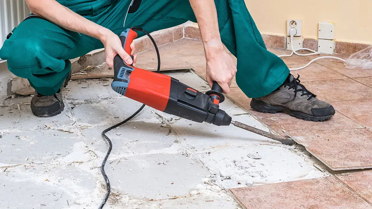A person in green overalls uses a jackhammer to remove ceramic tile flooring, breaking up the old tiles and concrete.