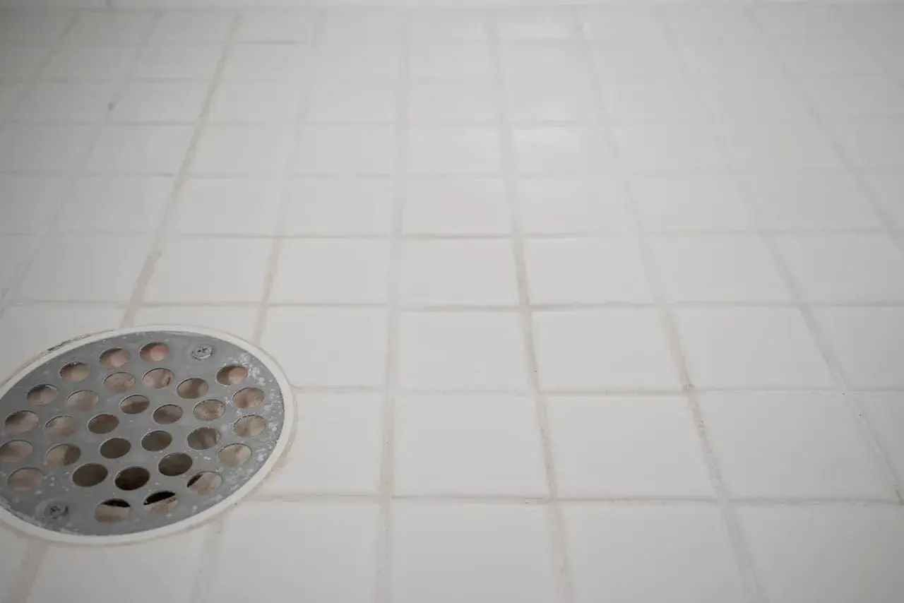 White tiled shower floor with a metal drain cover. The grout lines show signs of wear, indicating a need for tile grout repair.