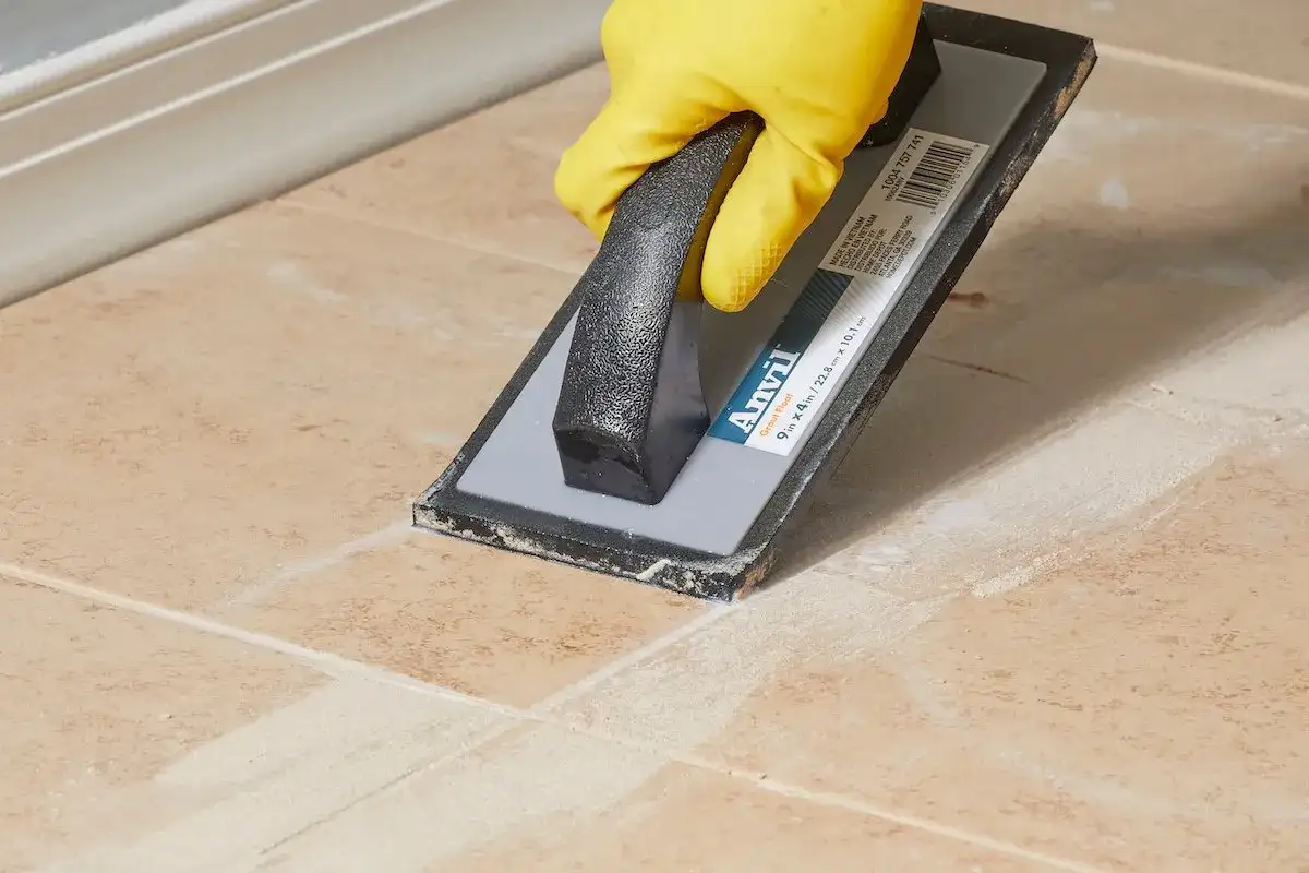 Yellow-gloved hand uses a grout float to remove old grout from tan tiles, demonstrating how to remove grout from tile.