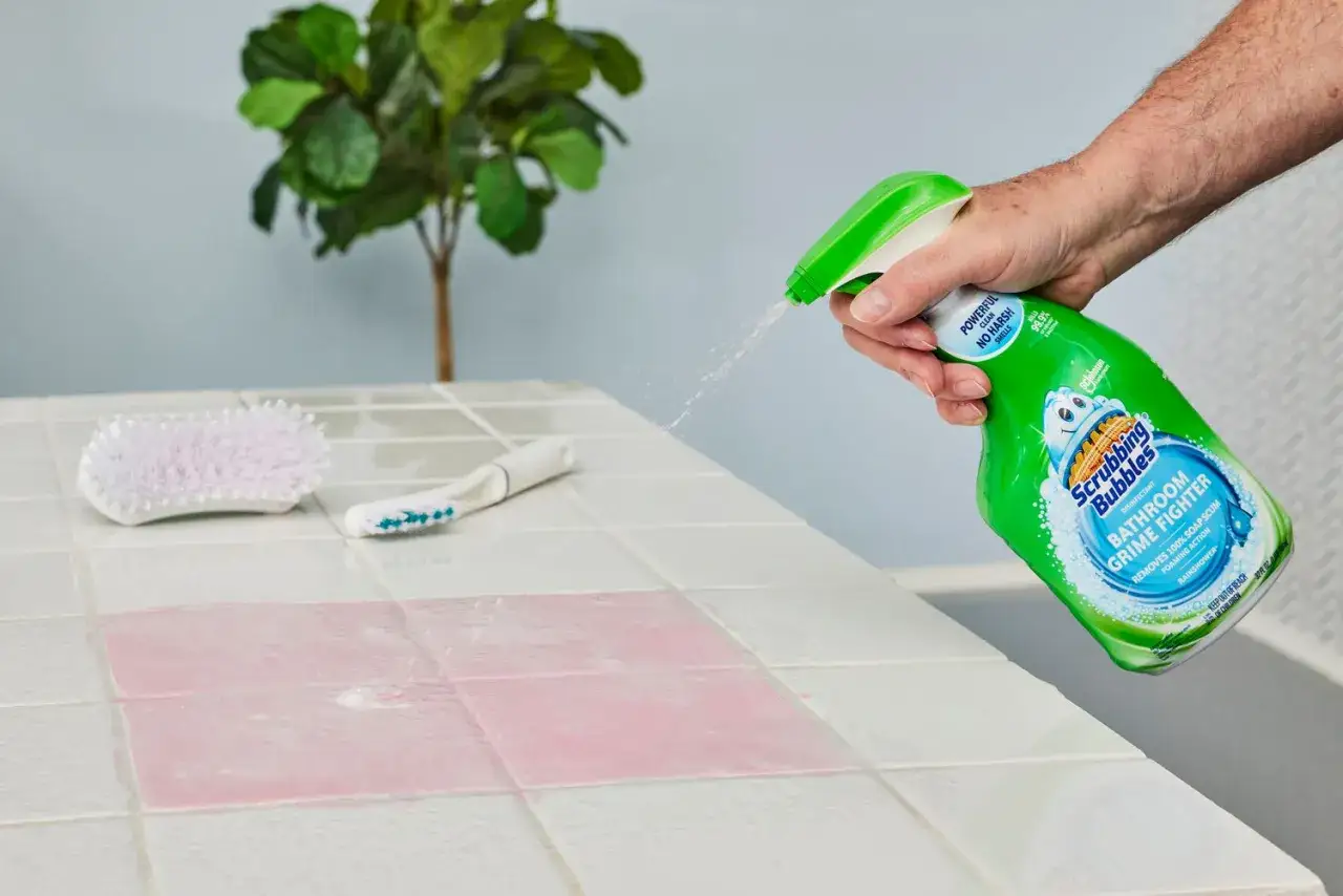 A hand sprays Scrubbing Bubbles Bathroom Grime Fighter onto tiled shower walls, showing how to clean grout in shower with a pink stain and brushes nearby.