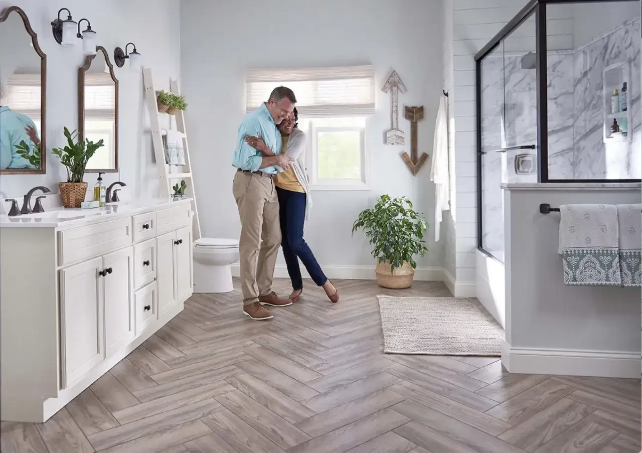 A couple embraces on a bathroom floor with herringbone wood-look tiles, showcasing the best flooring for kitchens and bathrooms.
