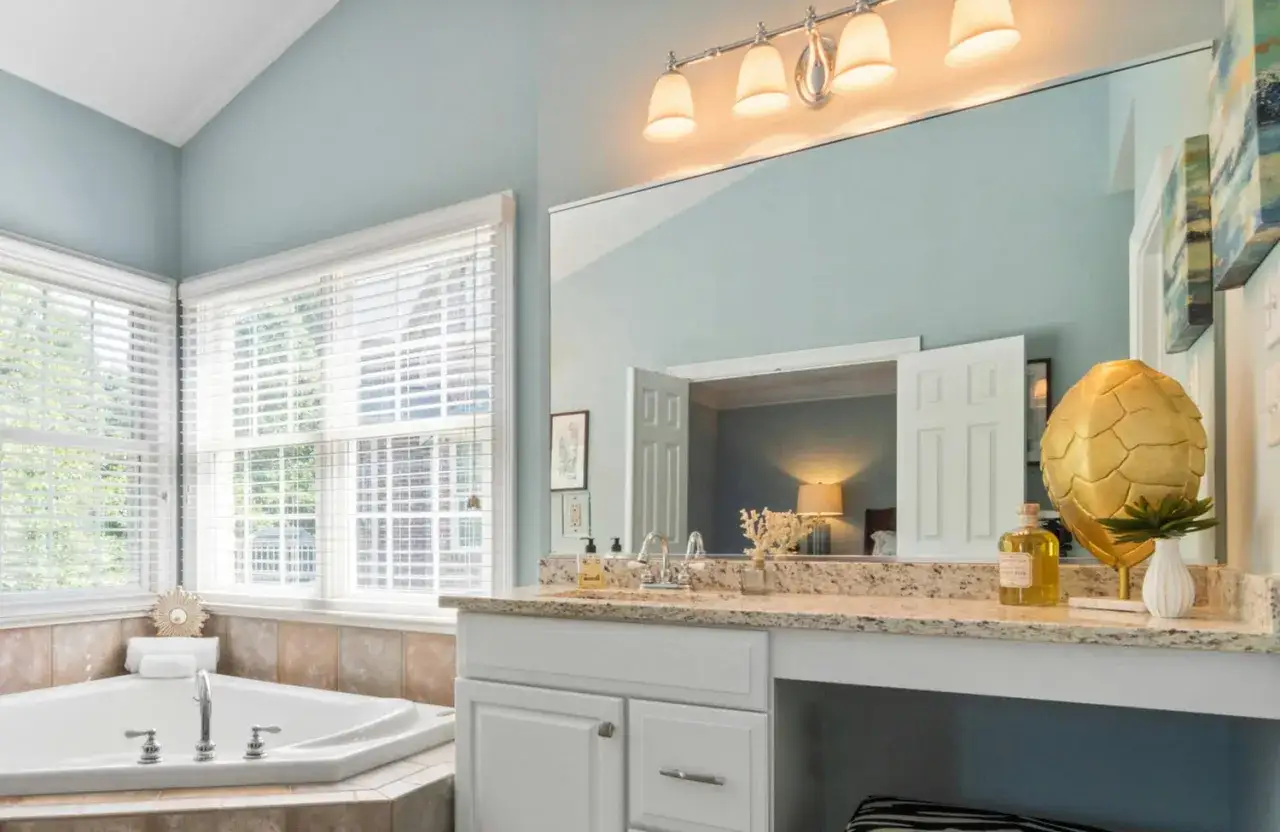 A bright bathroom with a corner tub and vanity. A large mirror above the vanity amplifies the light, using mirrors to reflect light from the windows and overhead fixtures.