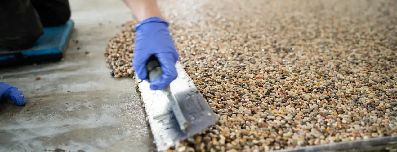 A gloved hand uses a trowel to smooth a textured surface during stone flooring installation, creating a beautiful pebble finish.