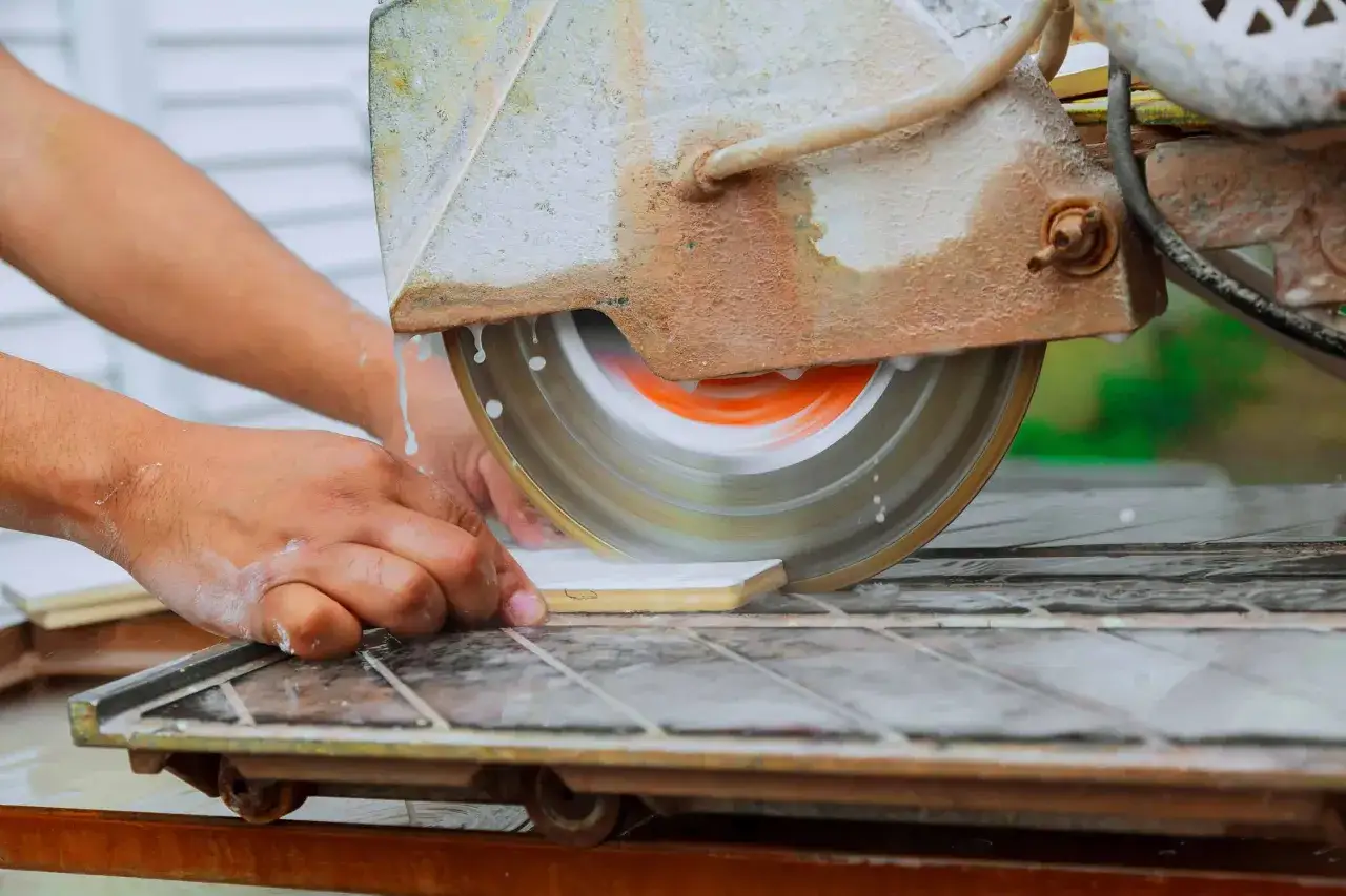 Hands guide a tile on a wet saw, demonstrating how to use a wet saw for precise cuts. Water sprays as the blade spins.