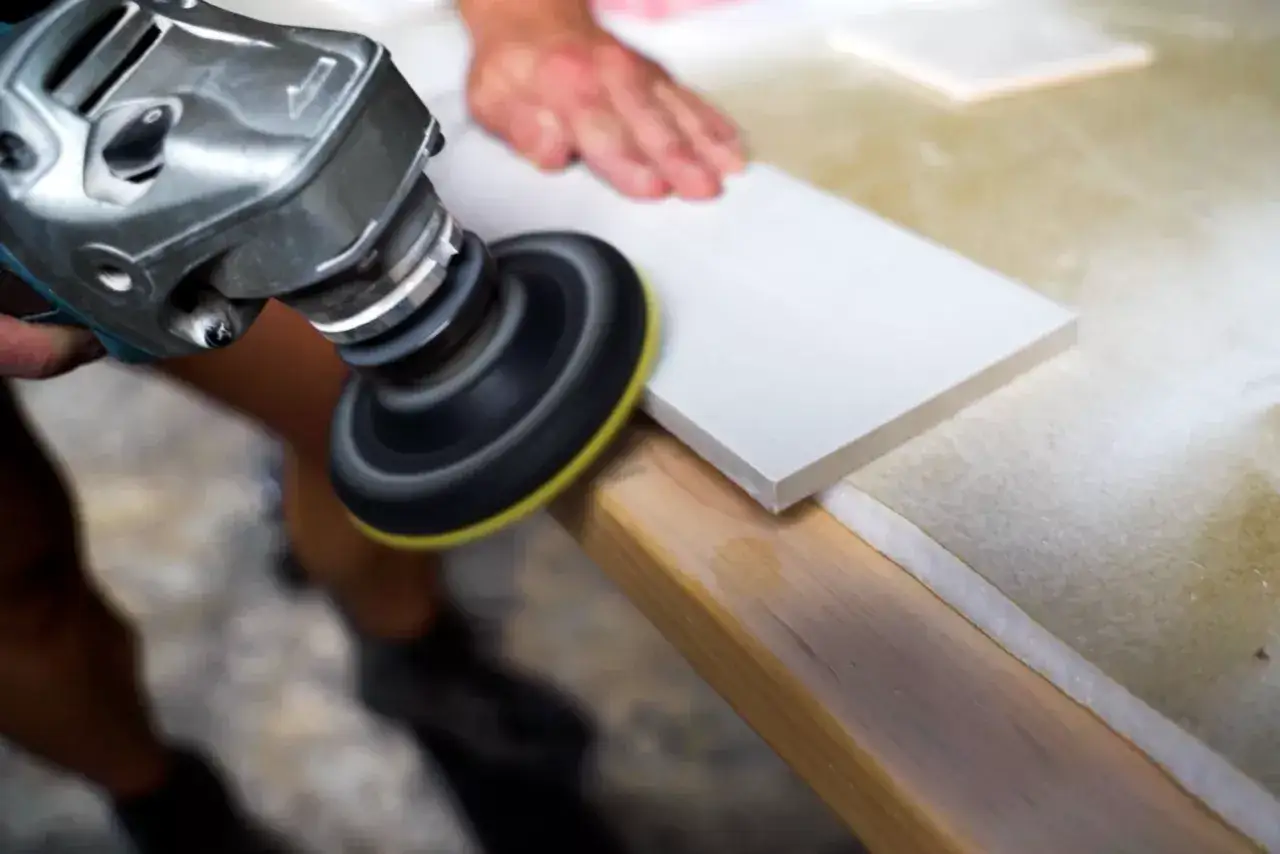 A craftsman uses an angle grinder to smooth the edge of a white cut stone tile.