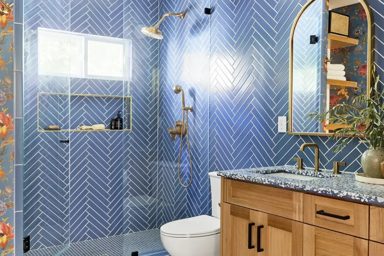 A modern bathroom featuring a stunning blue herringbone pattern shower, a wooden vanity, and a gold-framed mirror.