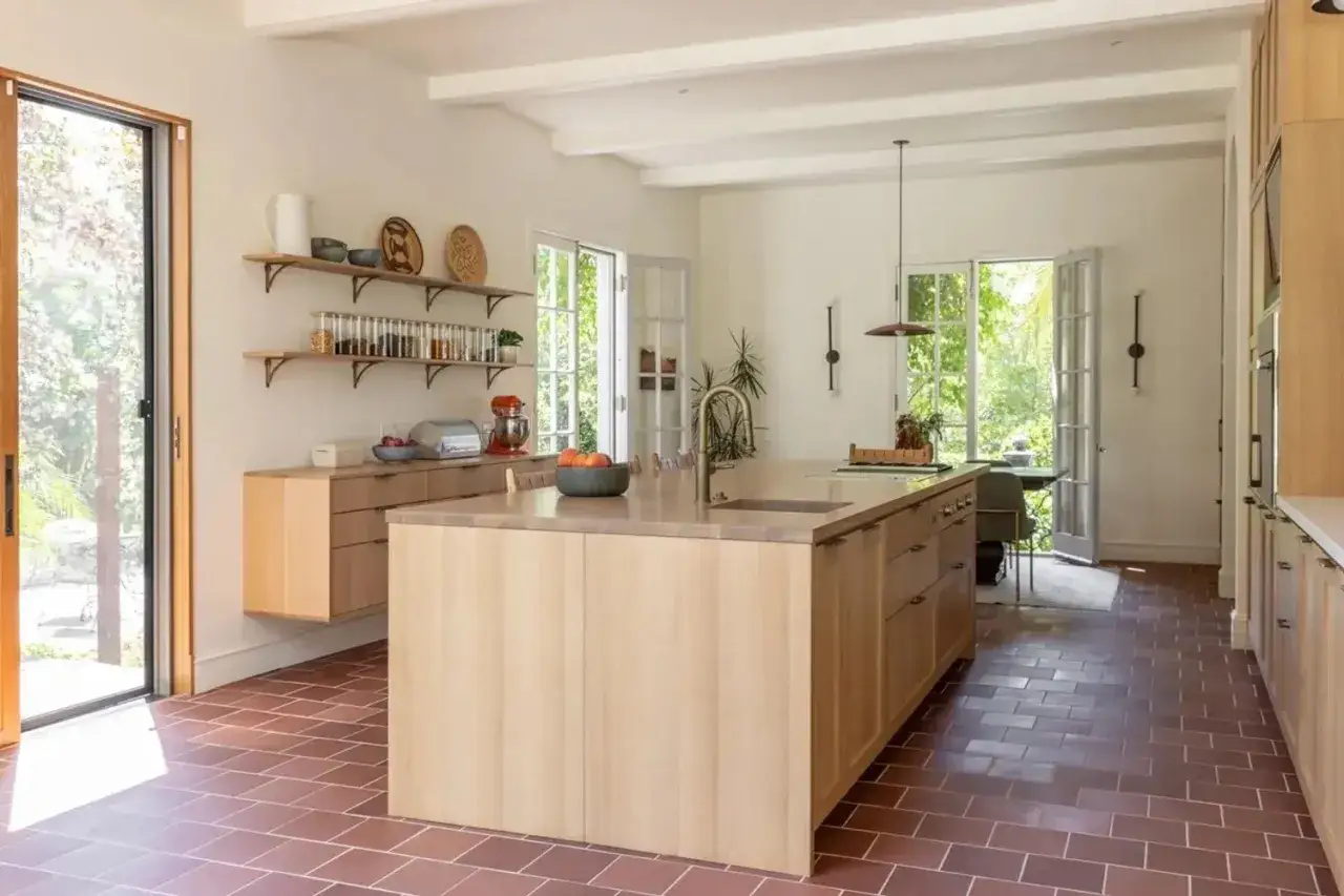 A modern kitchen with light wood cabinets and a large island, featuring warm terracotta tiling floors.