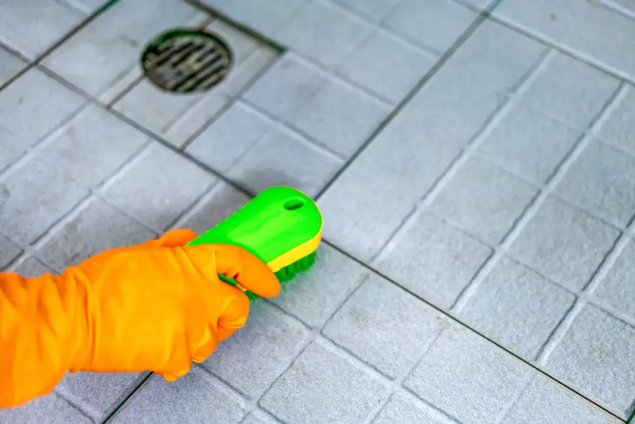 Orange-gloved hand scrubs grey ceramic tile with a green brush, demonstrating how to clean ceramic tile.