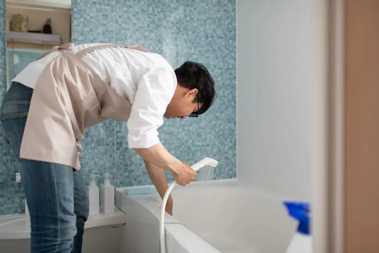 Man in apron rinses bathtub with showerhead, demonstrating how to clean bathtub.