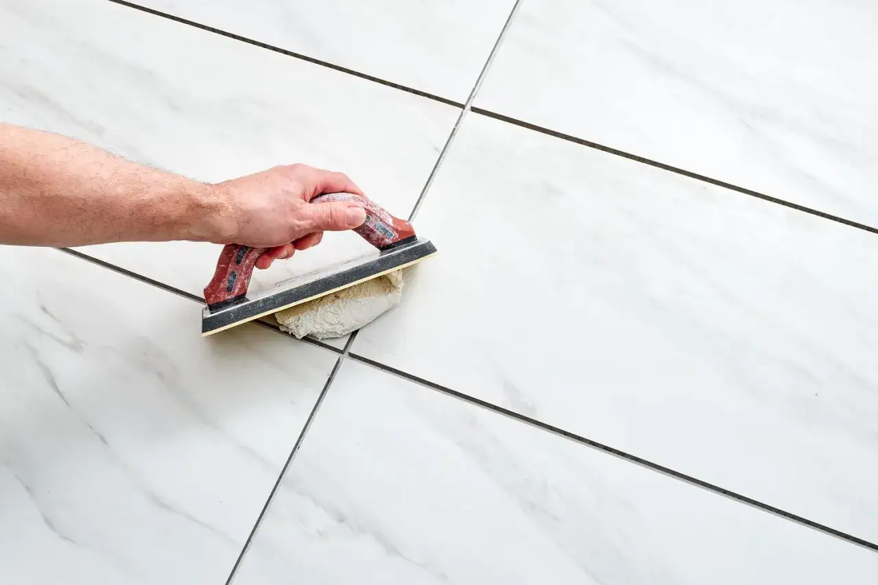 Hand applying grout to a white marble-patterned floor with a trowel.
