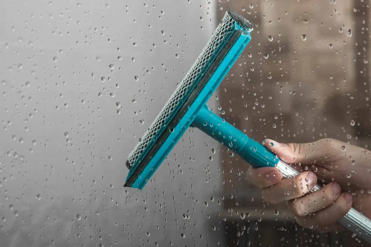 A hand uses a squeegee to clean water spots from shower glass, demonstrating how to clean shower glass for a streak-free shine.