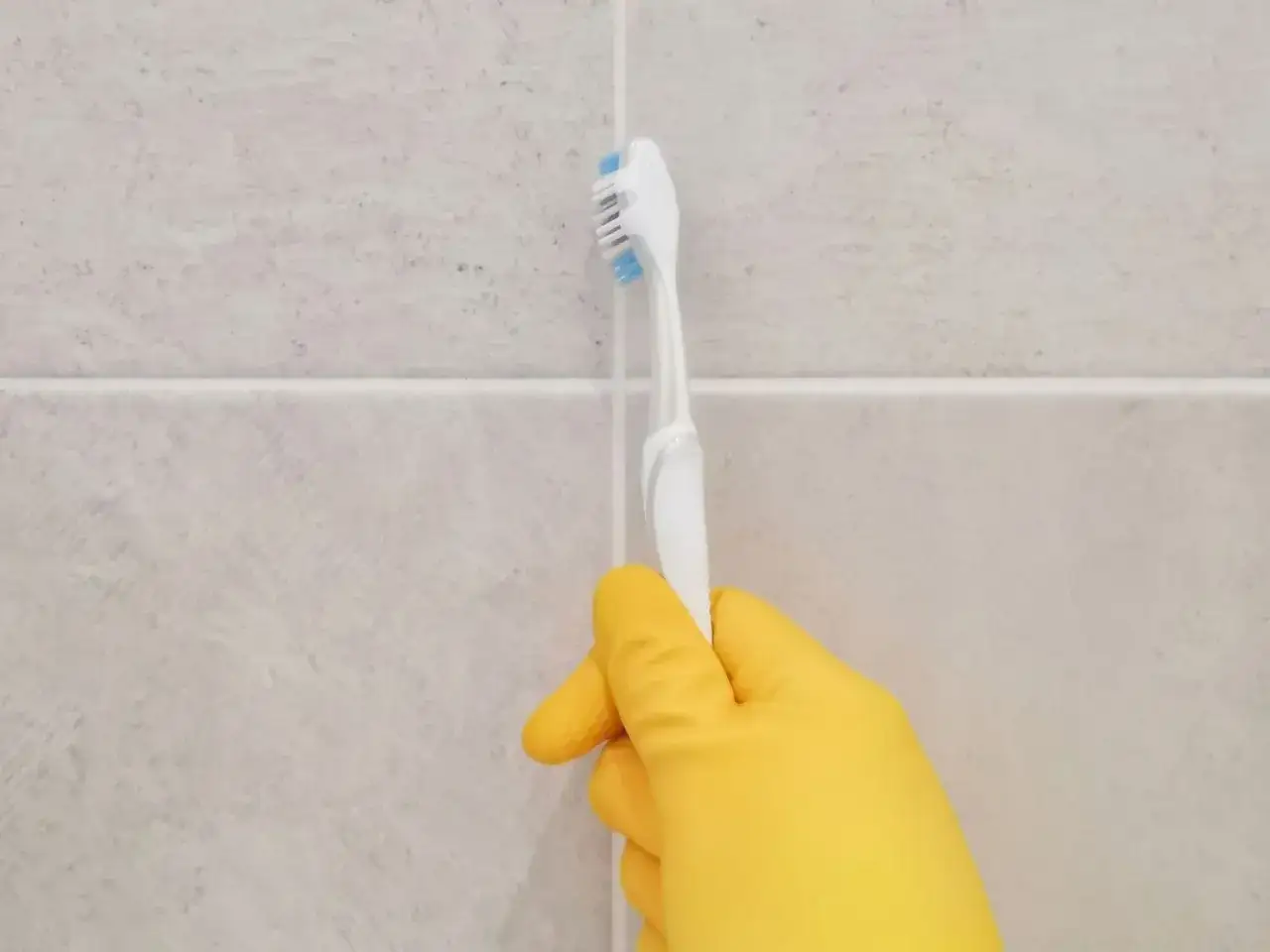 Yellow-gloved hand holding a toothbrush, demonstrating the best way to clean tile grout.