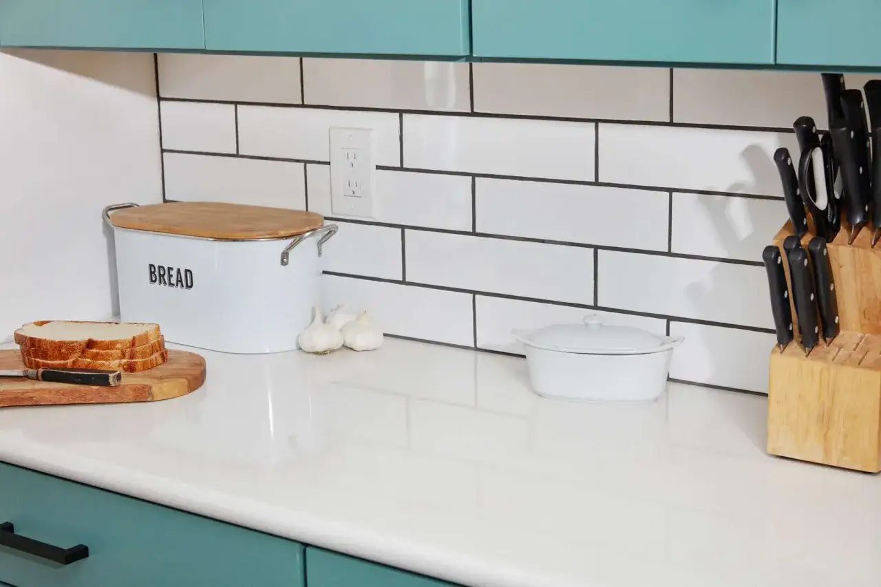 A kitchen counter with a bread box, sliced bread, garlic, a small pot, and a knife block against a random subway tile pattern backsplash.