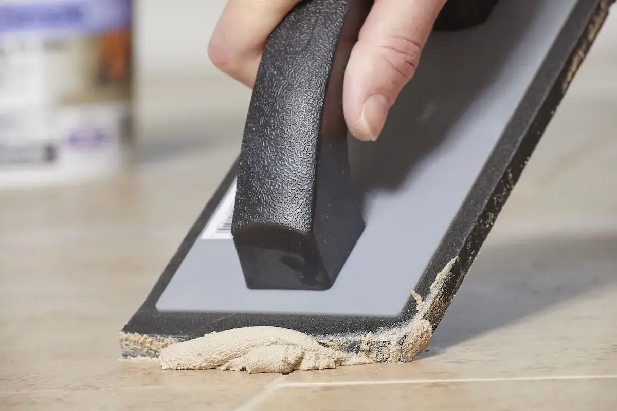 A hand uses a grout float to spread grout on tiles. It's normal for grout to crack if not applied correctly.