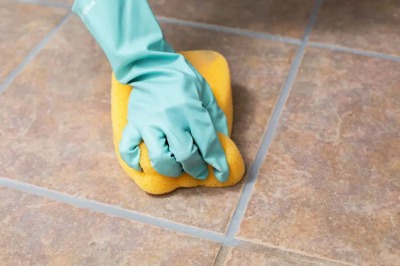 A gloved hand scrubs unglazed ceramic tile with a yellow sponge, demonstrating how to clean the porous surface.