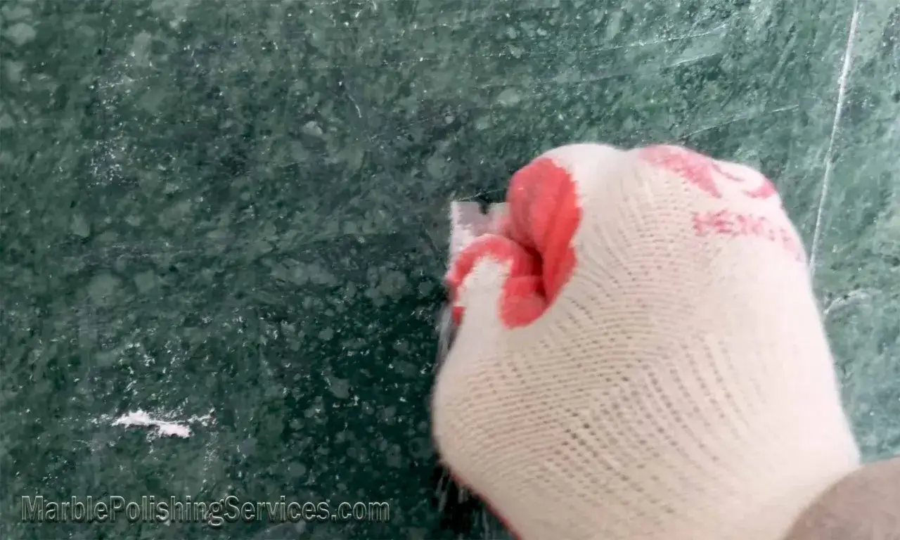 Gloved hand scraping white residue from green marble, demonstrating how to remove soap scum from marble.