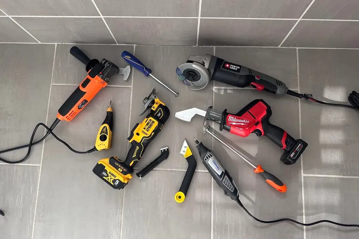Tools for tile grout repair: oscillating multi-tool, angle grinder, reciprocating saw, Dremel, and scrapers laid out on a tiled floor.