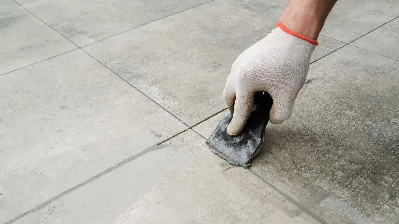Gloved hand uses a tool to apply grout to the seams between peel and stick tiles, completing the installation.