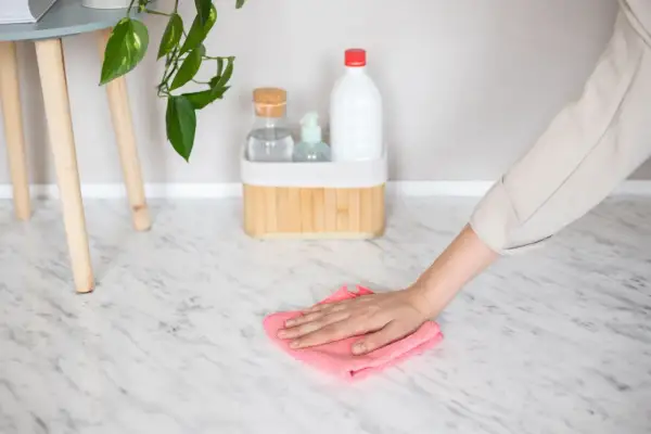 Hand wiping a marble floor with a pink cloth, with cleaning supplies in the background.
