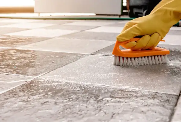 A gloved hand scrubs a tiled floor with a brush, showing the best way to clean porcelain tile.