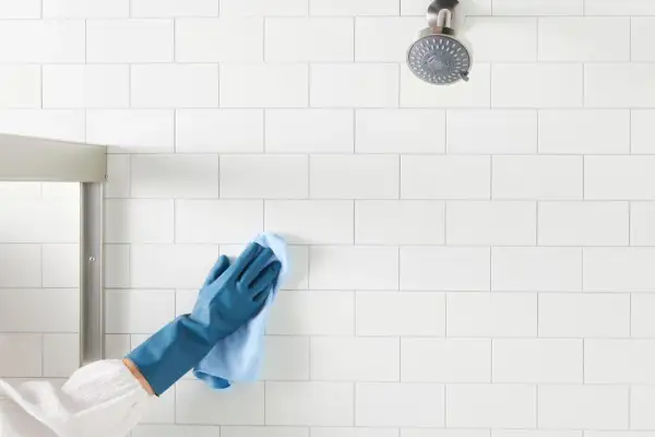 Gloved hand wipes white shower tiles, demonstrating how to clean shower tiles for a sparkling finish.