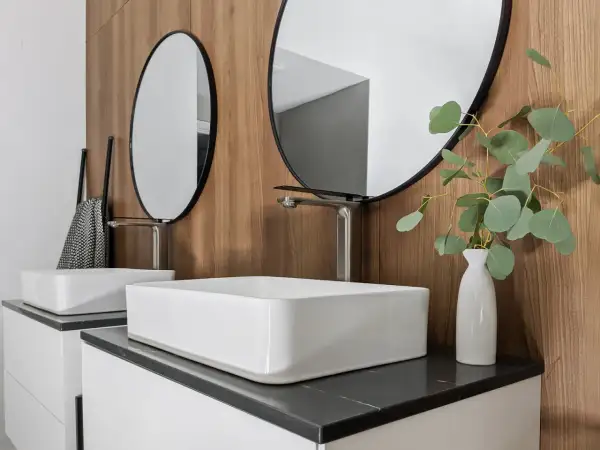 Modern bathroom with two white vessel sinks, sleek faucets, round mirrors, and a eucalyptus branch in a vase.