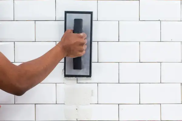 A hand applies grout to white subway tiles. The difference between sanded grout vs unsanded grout is visible in the texture.
