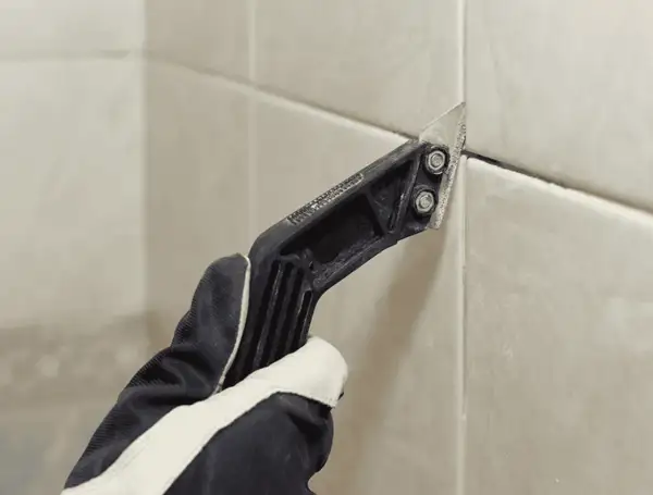 Gloved hand uses a grout removal tool to scrape away old grout, preparing for new grout over old grout.