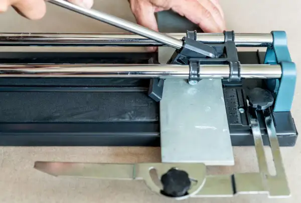 Hands guide a scoring wheel on a tile cutter, demonstrating how to cut ceramic tile by hand for a precise DIY project.