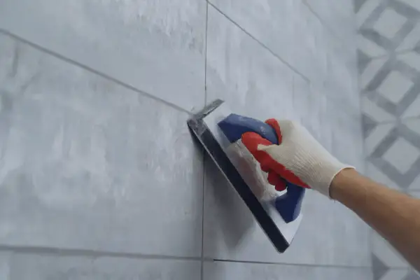 Hand in glove using a trowel for repairing grouting between light grey tiles.