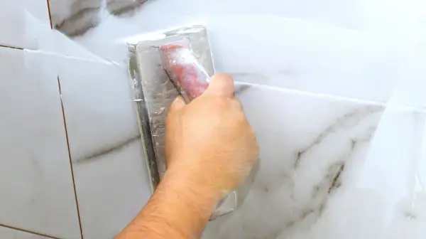 A hand uses a grout float to apply white grout to marble tiles. The grout is still wet, so it's too early to know how long does it take for grout to set.