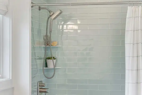 Modern shower with light blue subway tiles. A shower head and hose hang next to a small plant. This is the best way to regrout tiles for a fresh look.