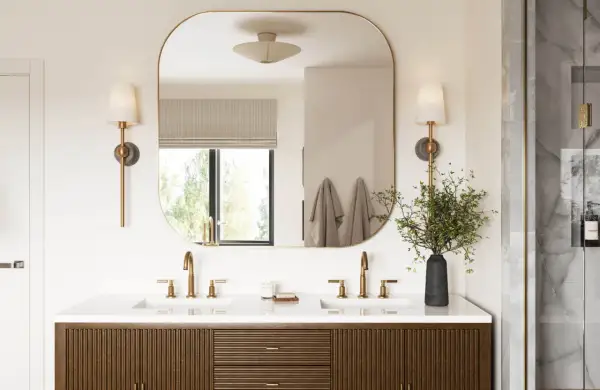 A modern bathroom vanity with dual sinks and brass fixtures. The large, rounded mirror reflects the window and wall sconces, effectively using mirrors to reflect light and brighten the space.