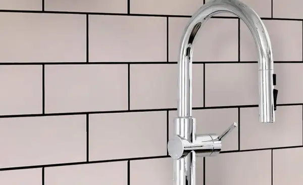 A modern chrome faucet stands against a backdrop of light pink subway tiles with smooth grout.