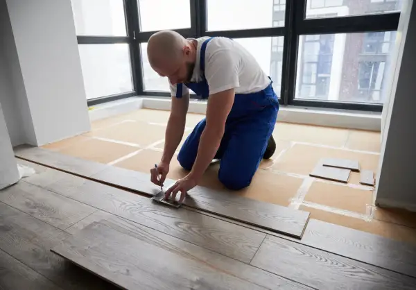 A man in blue overalls measures a laminate plank, demonstrating how to install laminate flooring.