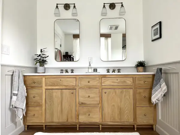 A beautiful DIY bathroom vanity with a natural wood finish, double sinks, and brass fixtures. Two framed mirrors hang above, reflecting the bright, airy space.