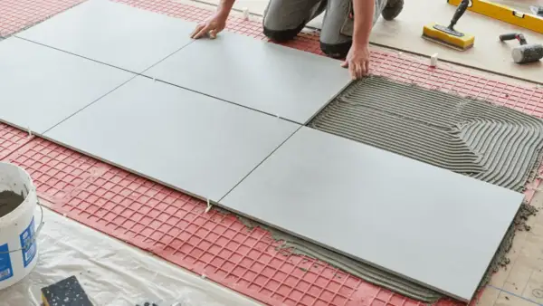 Tiling floors in a kitchen with large grey tiles and adhesive. Tools and a bucket of grout are nearby.