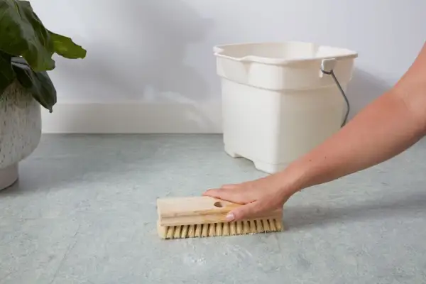 A hand scrubs a linoleum floor with a brush, with a bucket nearby. This shows how to clean linoleum floors effectively.