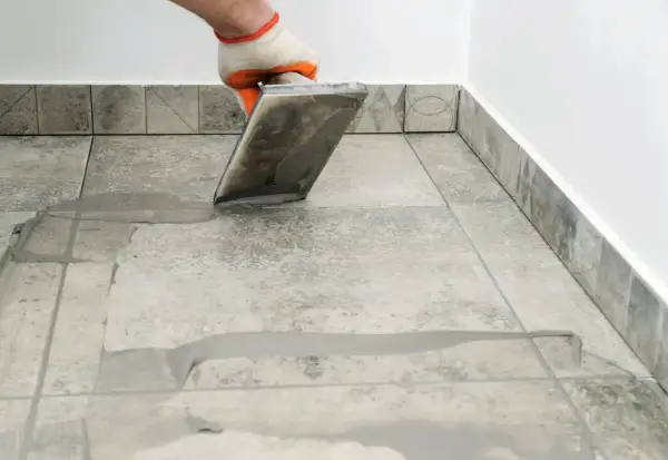 Gloved hand applies grout to tiled floor. Wondering how long for grout to dry? This process is key to a finished look.