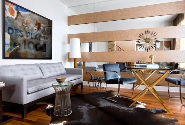 A large wall mirror with wood accents offers a stylish way to decorate around it. A grey sofa, abstract art, and a glass desk complete the modern living space.