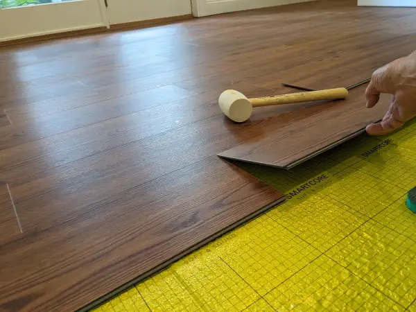 A hand is installing a dark wood-look LVT plank flooring over yellow underlayment. A rubber mallet rests nearby.