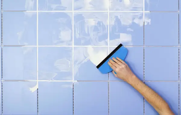 A hand uses a blue squeegee to spread white grout on light blue tiles, demonstrating how to clean tile grout.