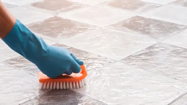 Gloved hand scrubs tile grout with an orange brush, demonstrating how to clean grout on a tile floor.