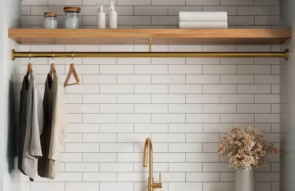 A laundry room with white tile beige grout walls, a wooden shelf, and a brass rod holding clothes.