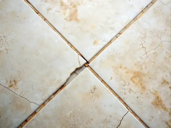 Cracked grout between beige floor tiles. Consider how often should you regrout floor tiles to prevent this damage and maintain your floor's integrity.