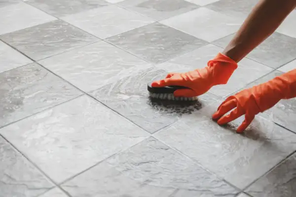 Hands in orange gloves scrub a tiled floor. The person is working to polish porcelain tile, making it shine.