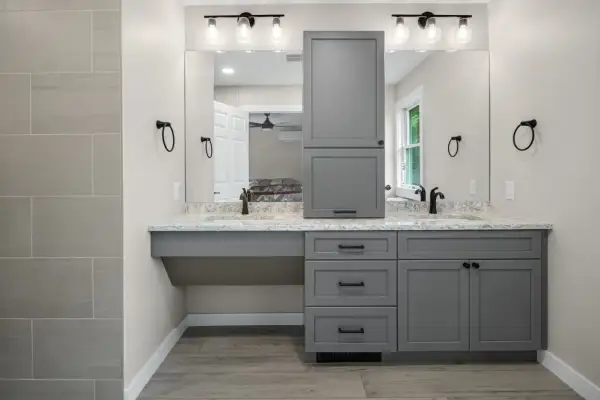 A modern, gray, freestanding vanity with dual sinks and a tall cabinet stands against a light gray wall.