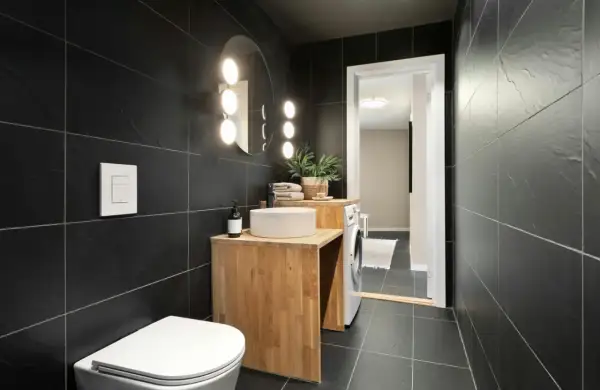 A modern bathroom with black tiles. Natural wood vanity, white sink, and toilet complement the dark tiles, showing what colour goes with black tiles in bathroom.