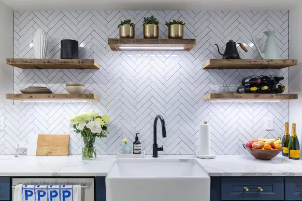A modern kitchen with a white chevron tile backsplash, rustic wood shelves, and a farmhouse sink. The chevron tile out of style? Not in this chic design!