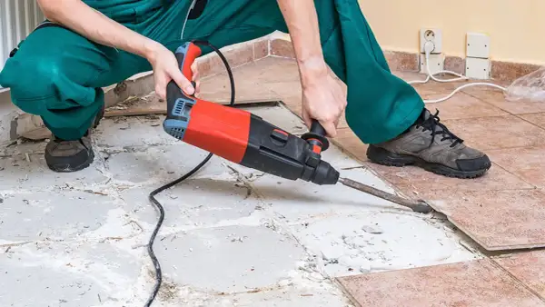 A person in green overalls uses a jackhammer to remove ceramic tile flooring, breaking up the old tiles and concrete.